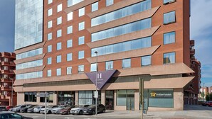 A multi-story hotel with a modern design, featuring a mix of brick and glass facade, and a visible parking area with several cars. - Apartamentos Hesperia Sant Joan (Sant Joan Despi)