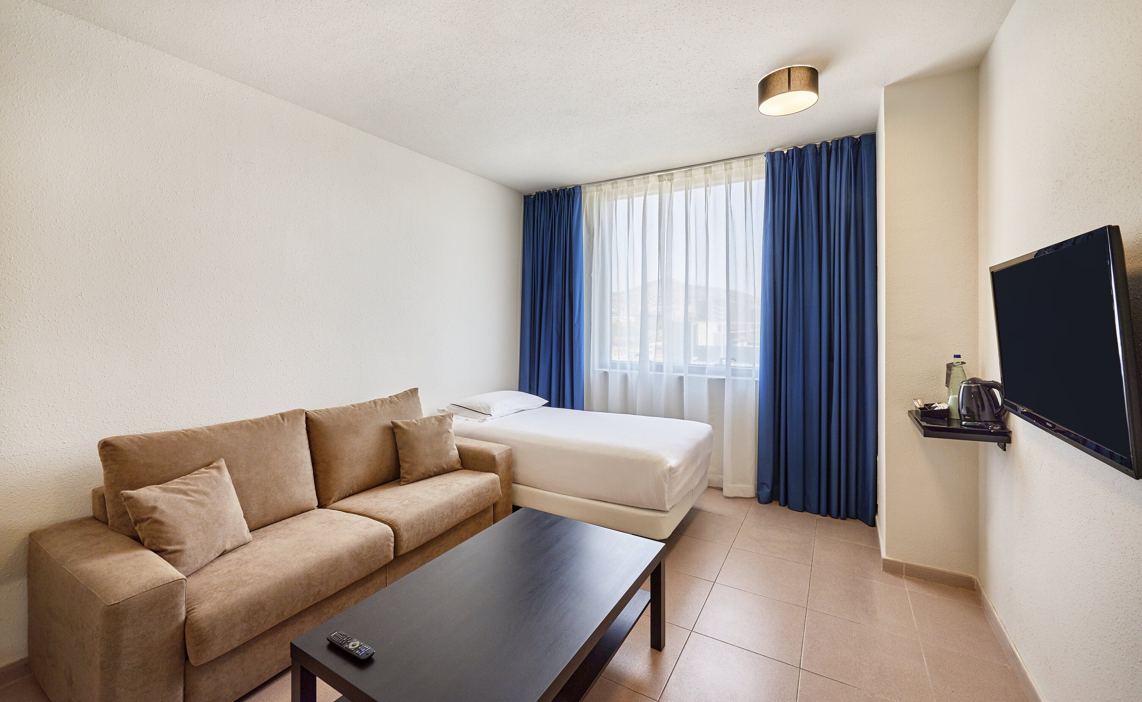 Habitació d'hotel moderna amb un sofà, una taula petita, un televisor i una finestra amb cortines.