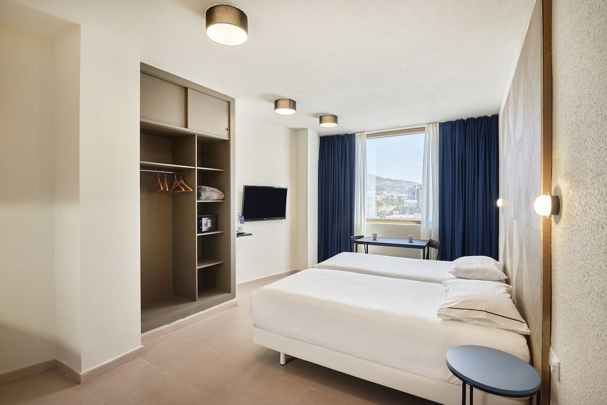 A hotel room with two beds, a wardrobe, a desk, a TV, and a window with curtains.