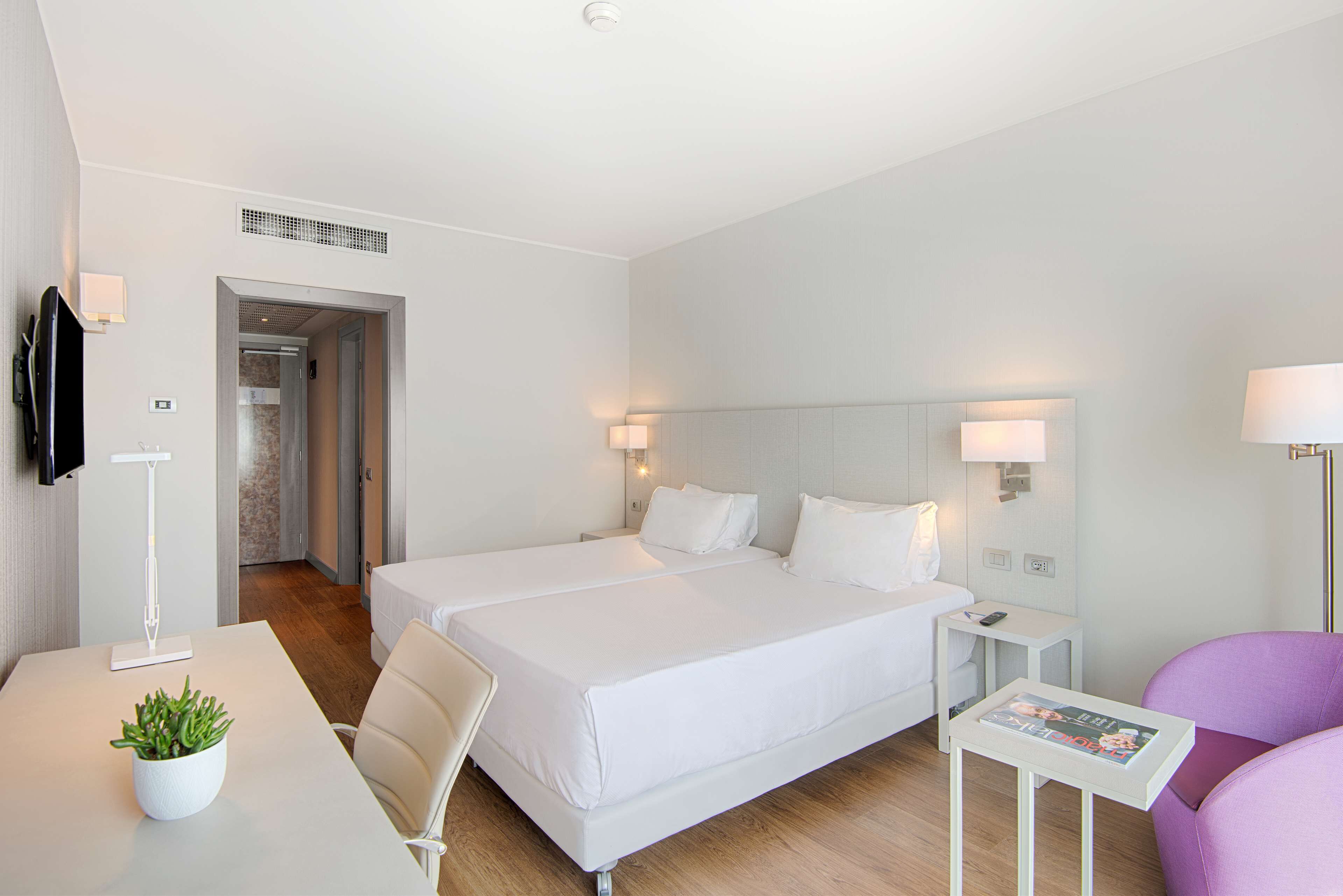 Habitación de hotel con una cama grande, un escritorio con una planta, una silla morada y una mesita con una revista.