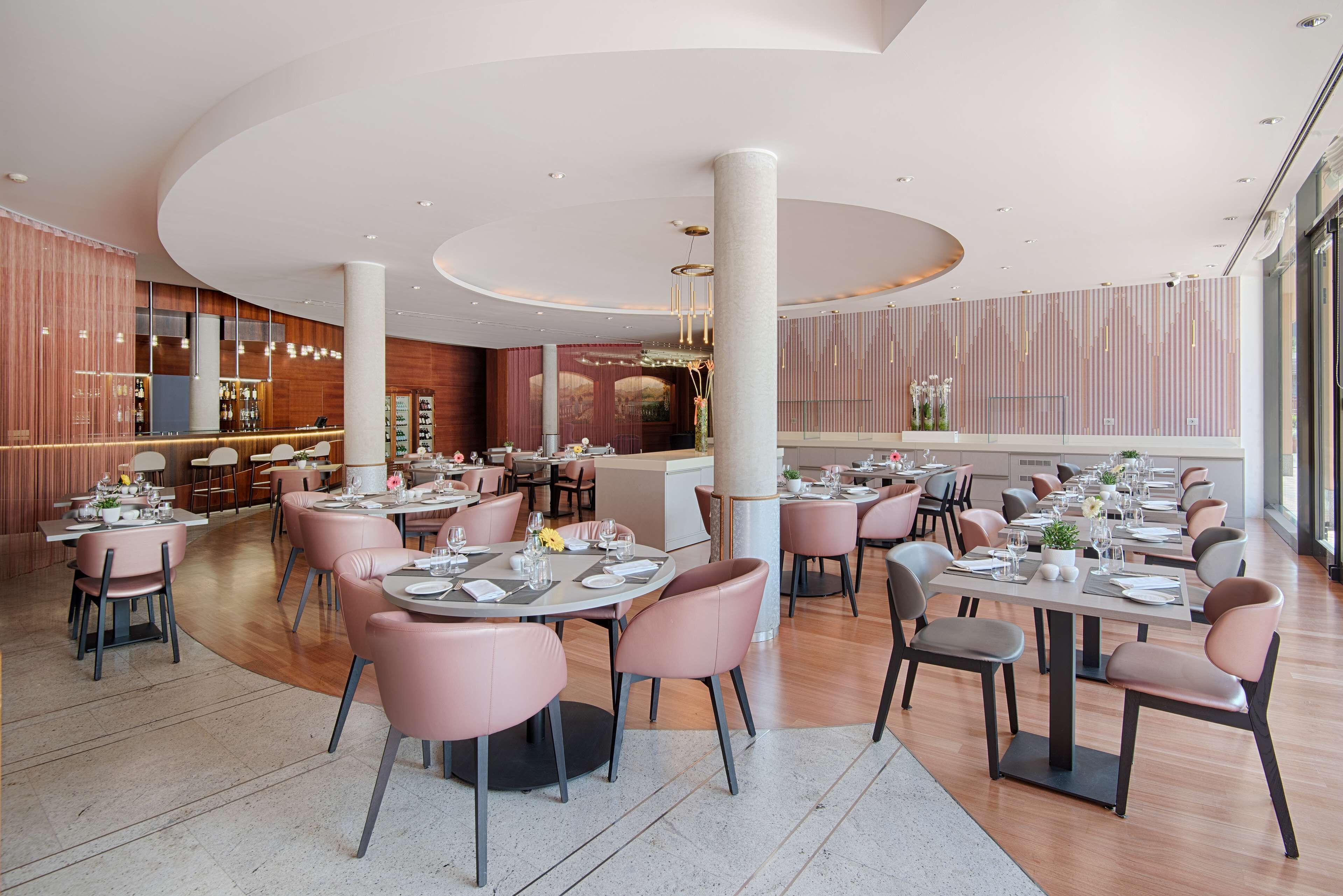 A modern restaurant with round tables and pink chairs, featuring a wooden interior and a central column.