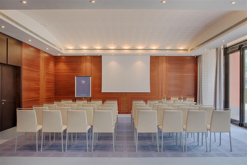 Uma sala de conferência com fileiras de cadeiras, uma tela de projetor e uma parede de painéis de madeira.