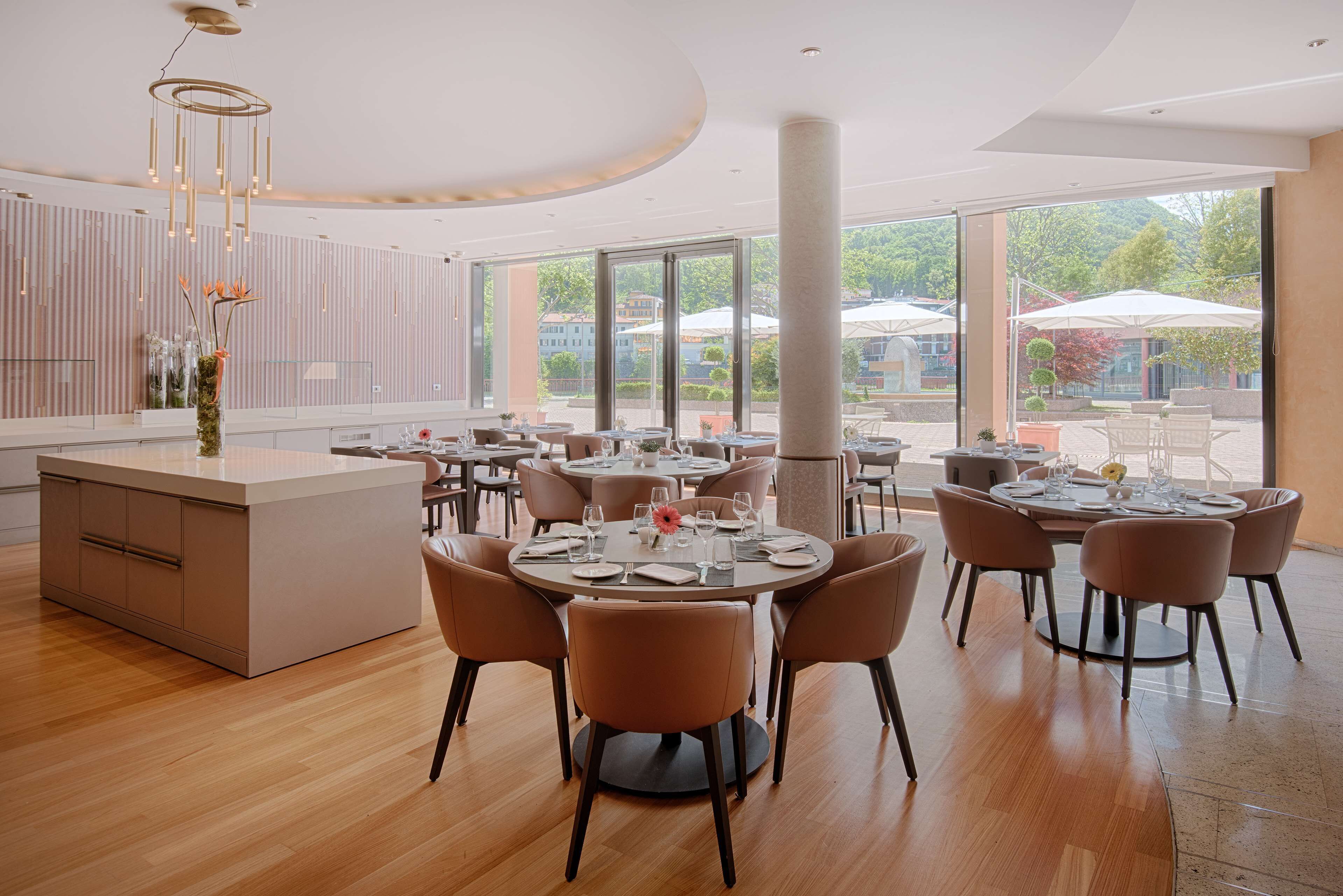 Un espace repas moderne avec des tables et des chaises rondes, un îlot central agrémenté d’un vase et de grandes fenêtres offrant une vue sur l’extérieur.