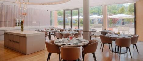 A modern dining area with round tables and chairs, a central island with a vase, and large windows offering a view of the outdoors.