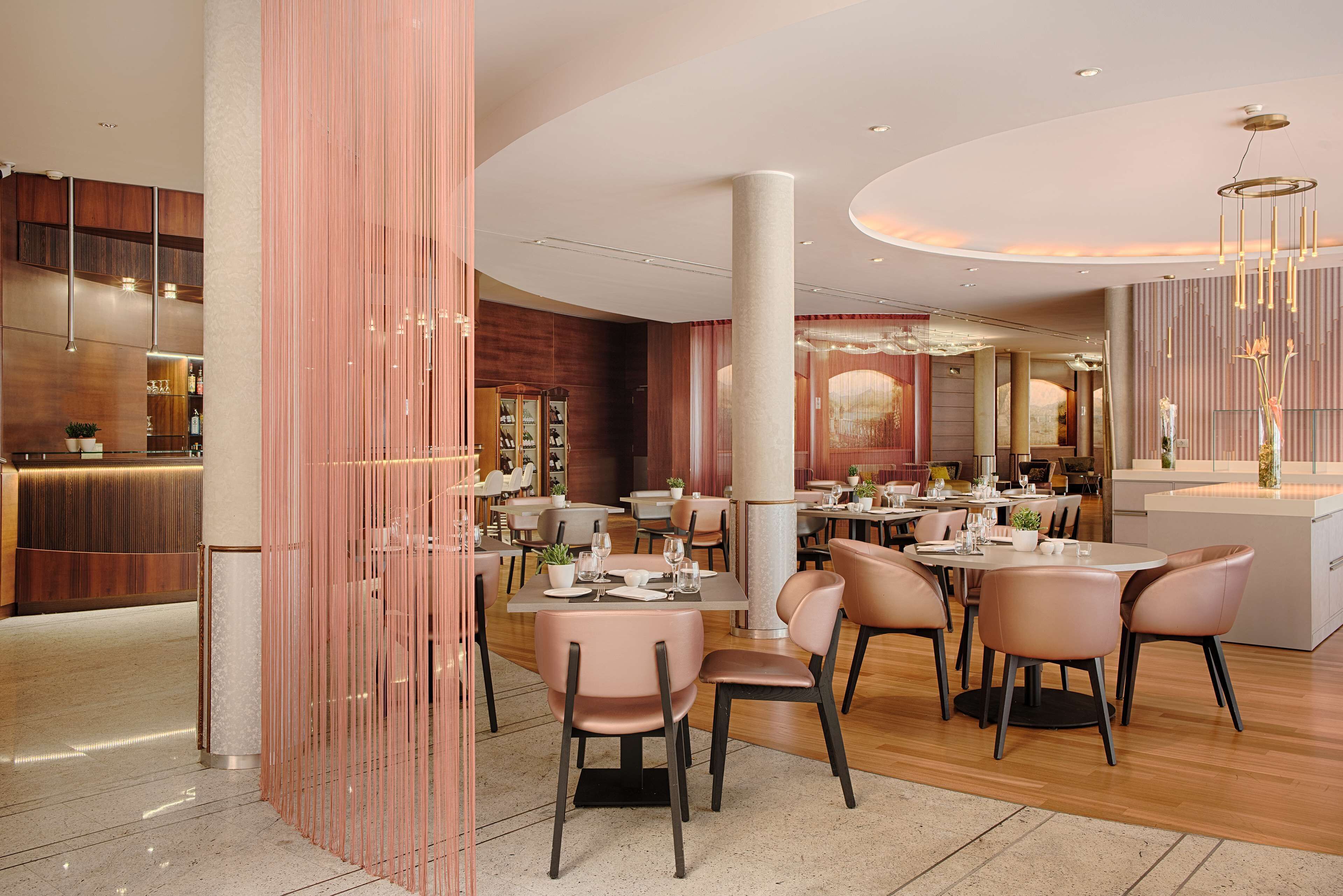 Een modern restaurant met ronde tafels en roze stoelen, een houten receptiebalie en een grote pilaar in het midden.