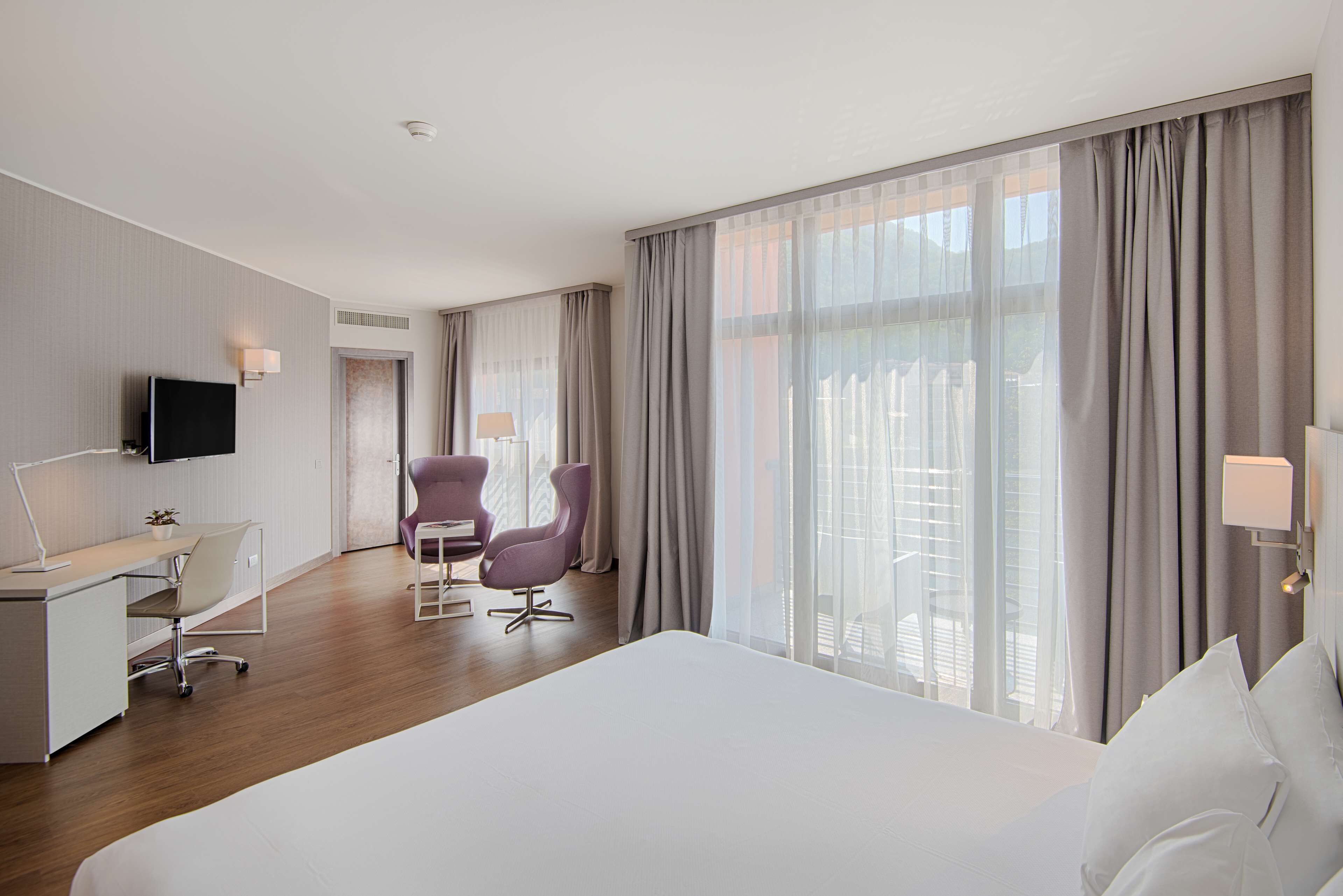 A hotel room with a large bed, a desk with a chair, a television, and a view of the outdoors through large windows.