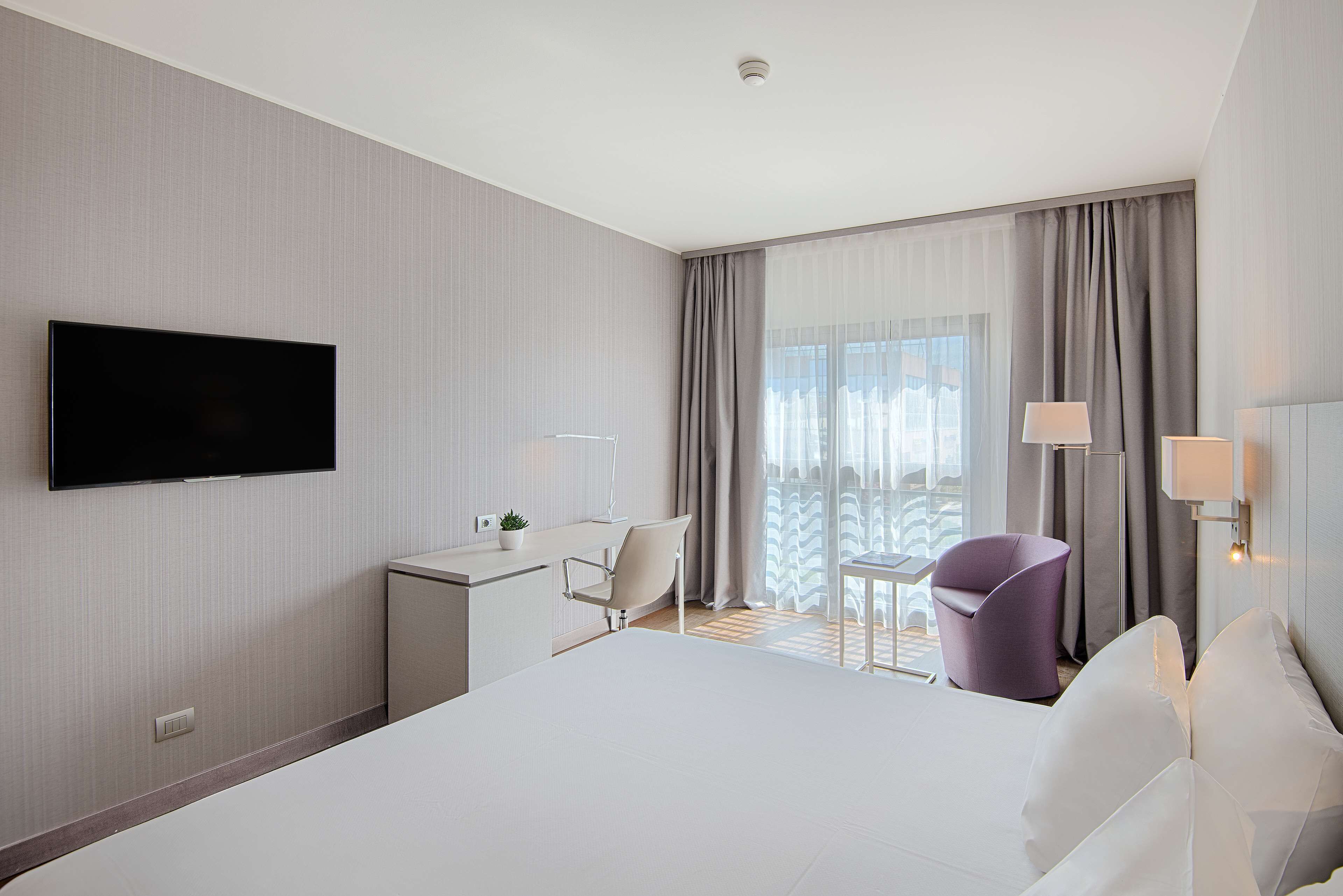 A modern hotel room with a large bed, a desk, a chair, and a TV mounted on the wall.