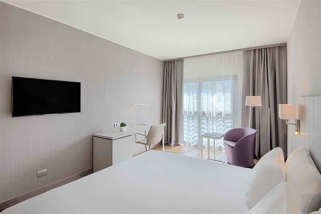 A modern hotel room with a large bed, a desk with a chair, a television mounted on the wall, and a balcony with curtains.