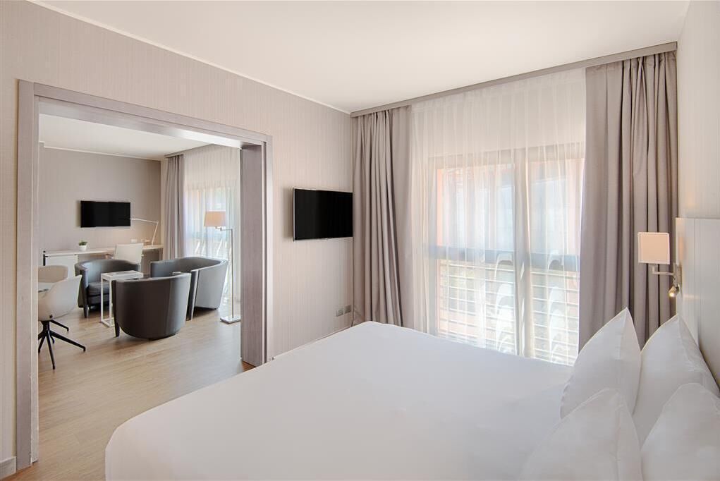 Una camera d'albergo con un letto grande, una scrivania con una sedia, una TV e un balcone con tende.