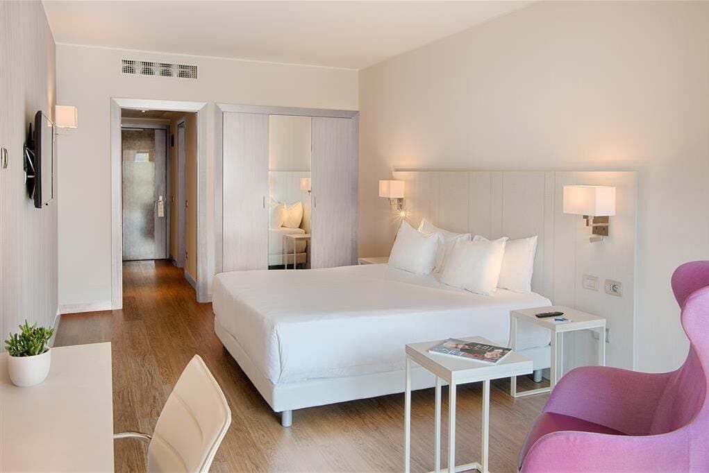 A modern hotel room with a large bed, bedside tables, a desk, a chair, and a pink chair.