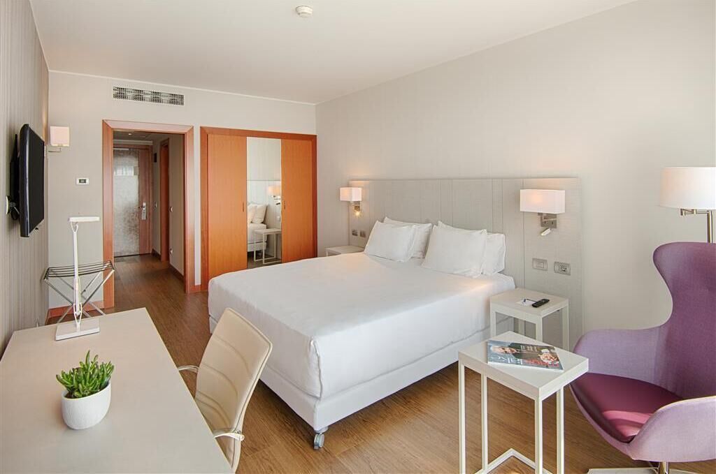 A hotel room with a large bed, a desk with a plant, a chair, a TV, and a lamp.