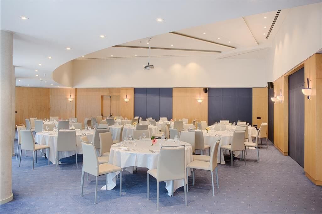 Un amplio salón de banquetes bien iluminado, con mesas y sillas redondas, equipado con proyectores instalados en el techo y apliques de pared.