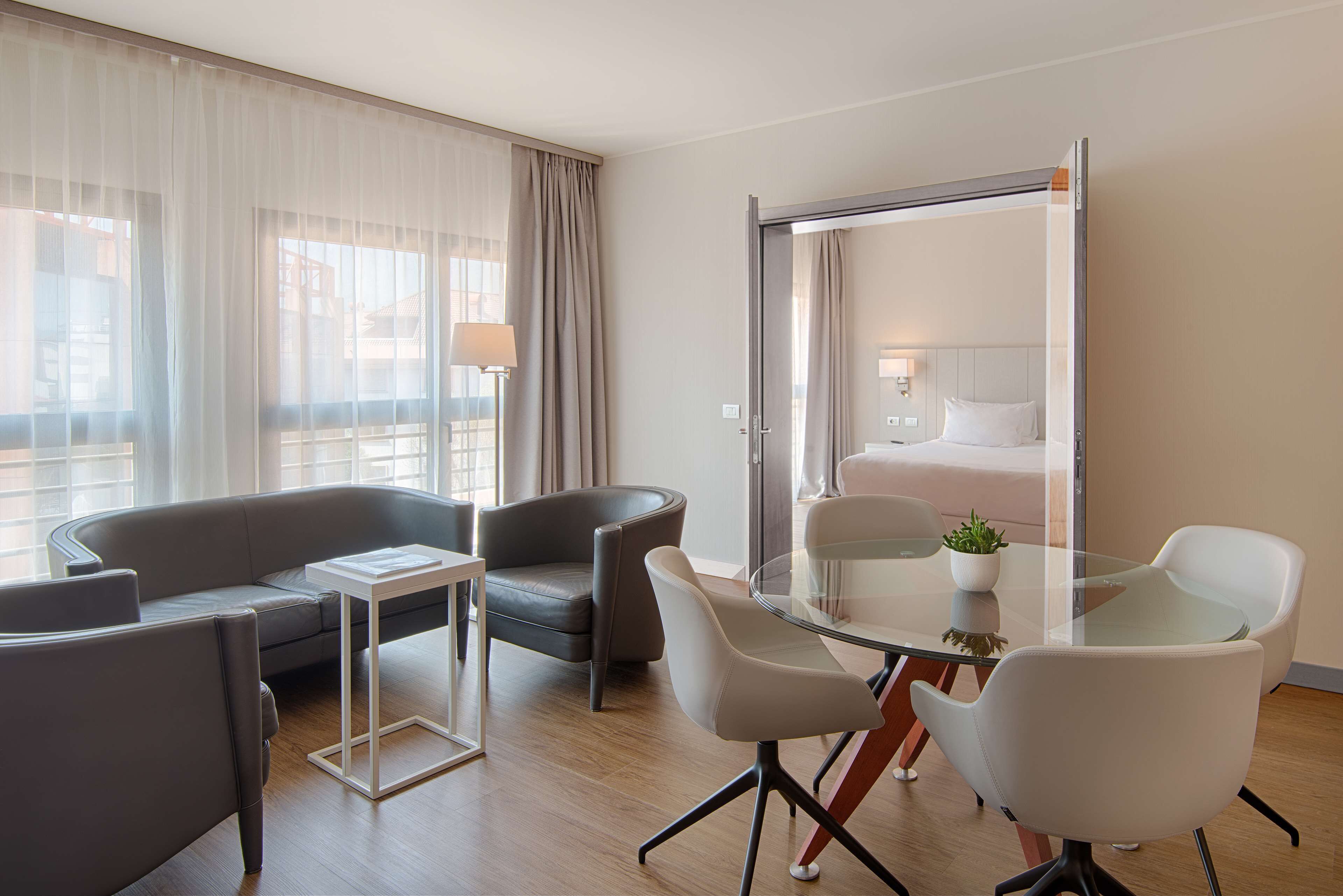Una habitación de hotel moderna con sofá, dos sillones, una mesa de vidrio y una cama con cabecera blanca.