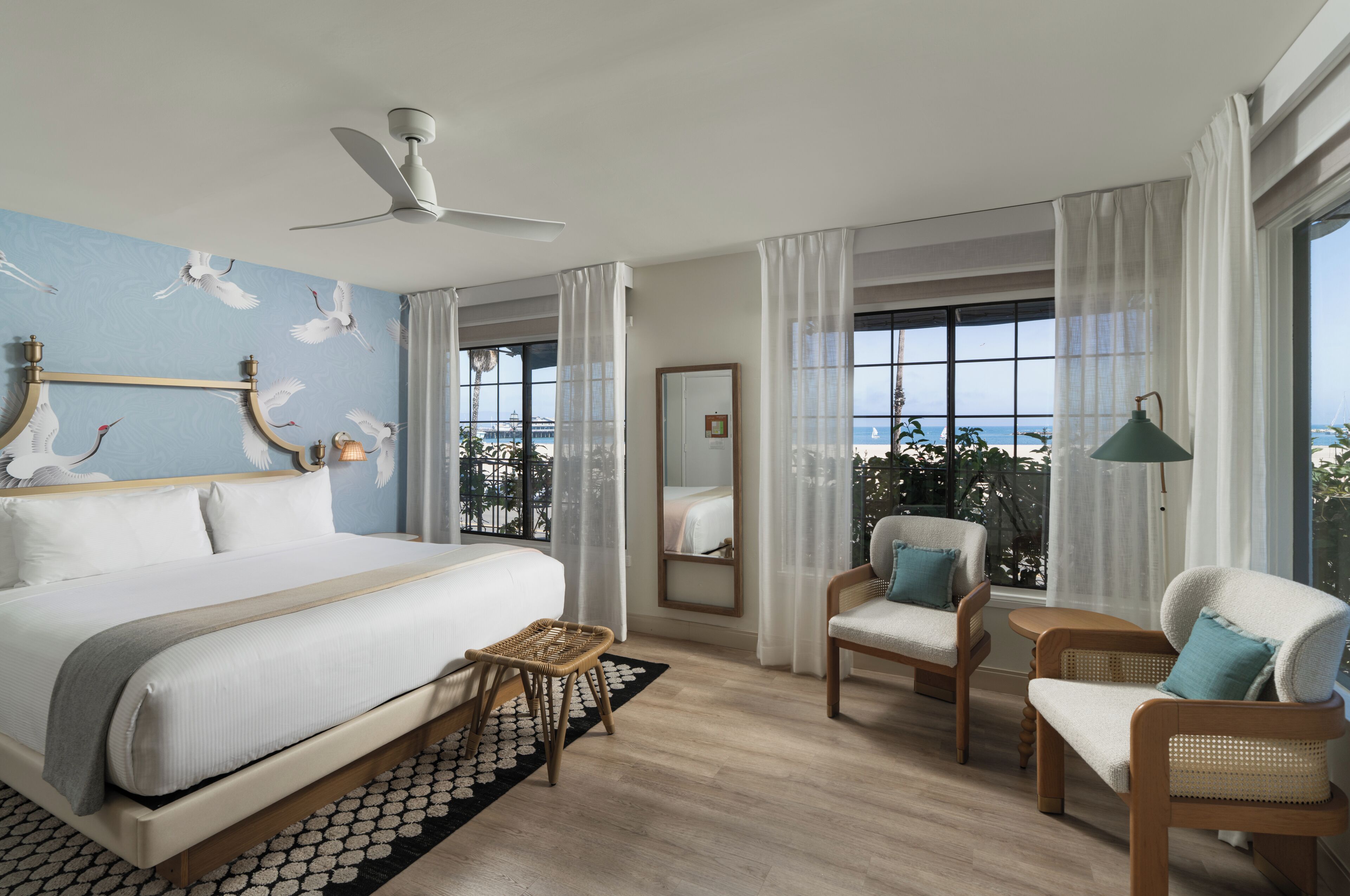 Quarto de hotel com cama, duas cadeiras, uma mesa pequena, ventilador de teto e uma janela grande com vista para a praia.