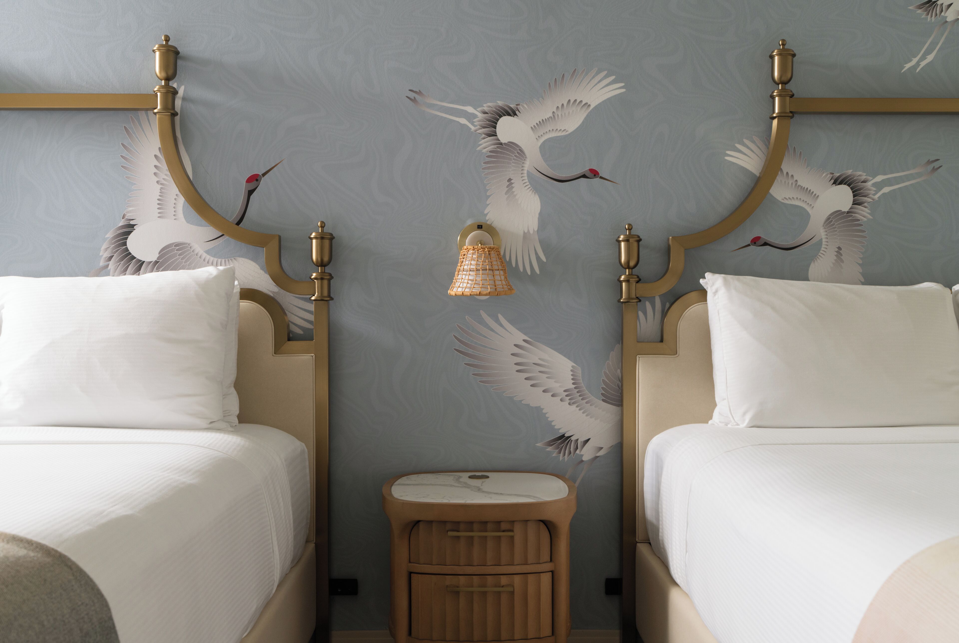 A room with two beds, a small wooden nightstand, and a wall with a bird mural.