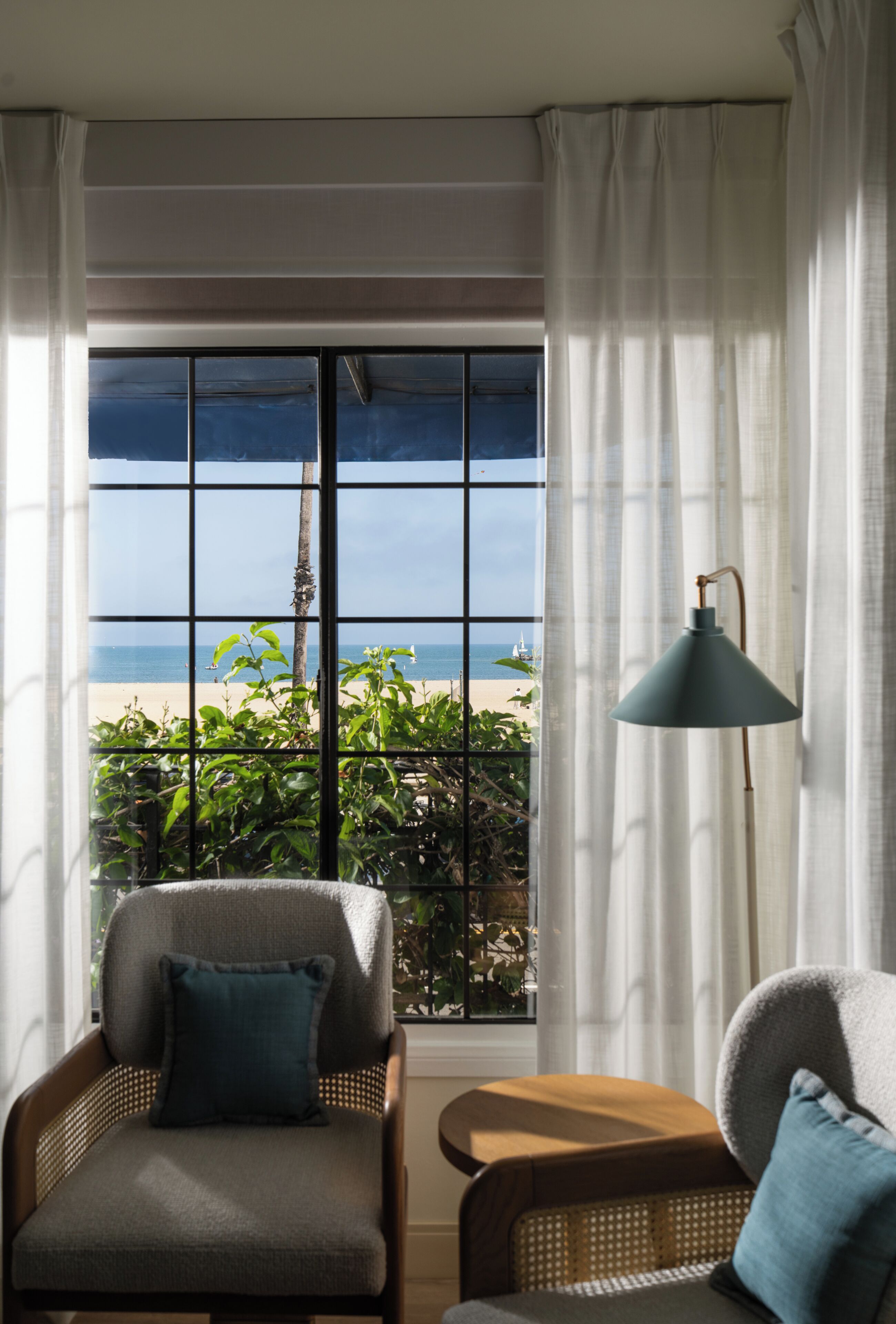 A room with a large window offering a view of the beach and ocean. There is a chair and a table with a lamp.