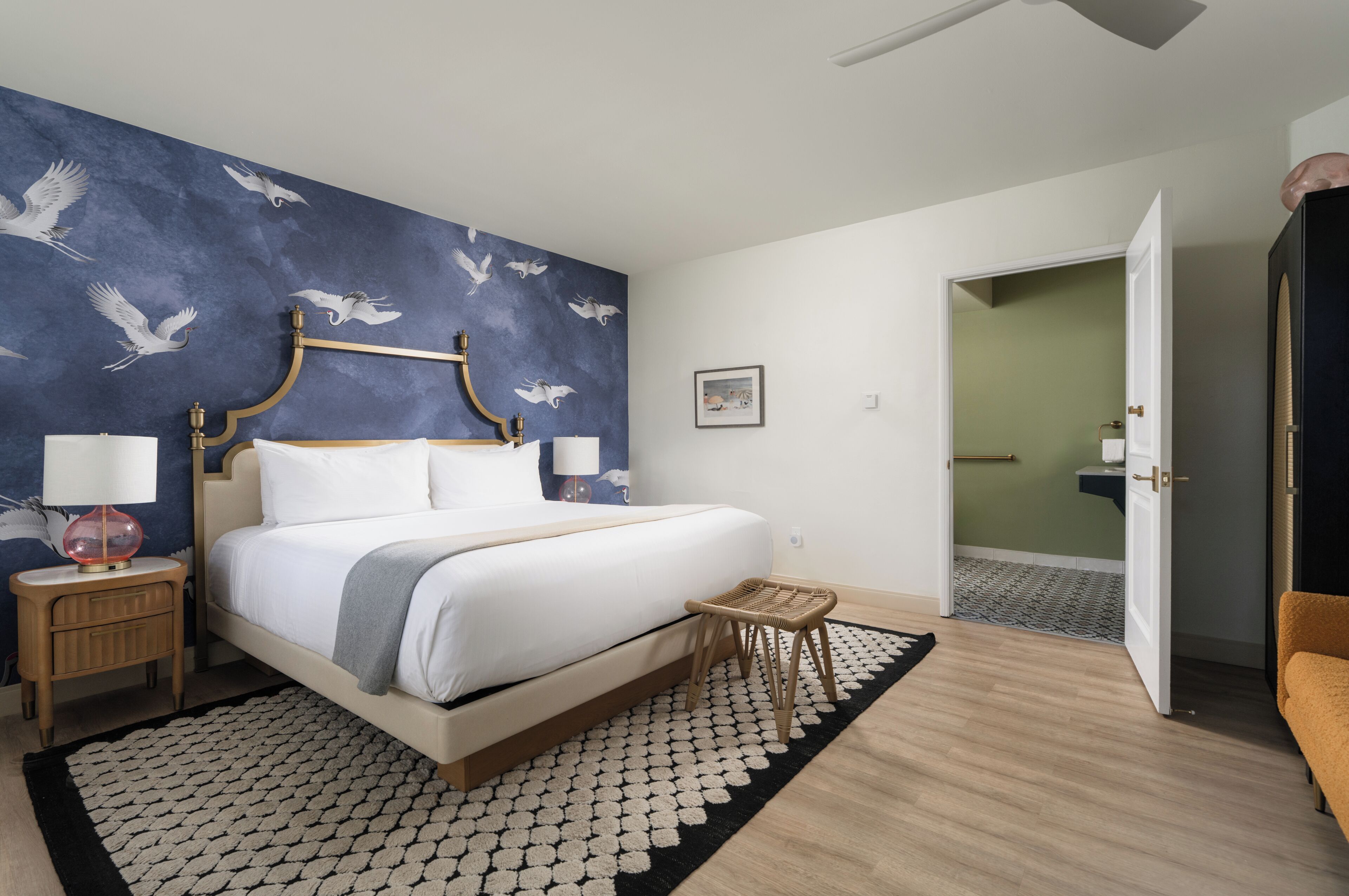A hotel room with a large bed, a nightstand, a bedside lamp, a small table, a wicker stool, and a wall with a bird mural.