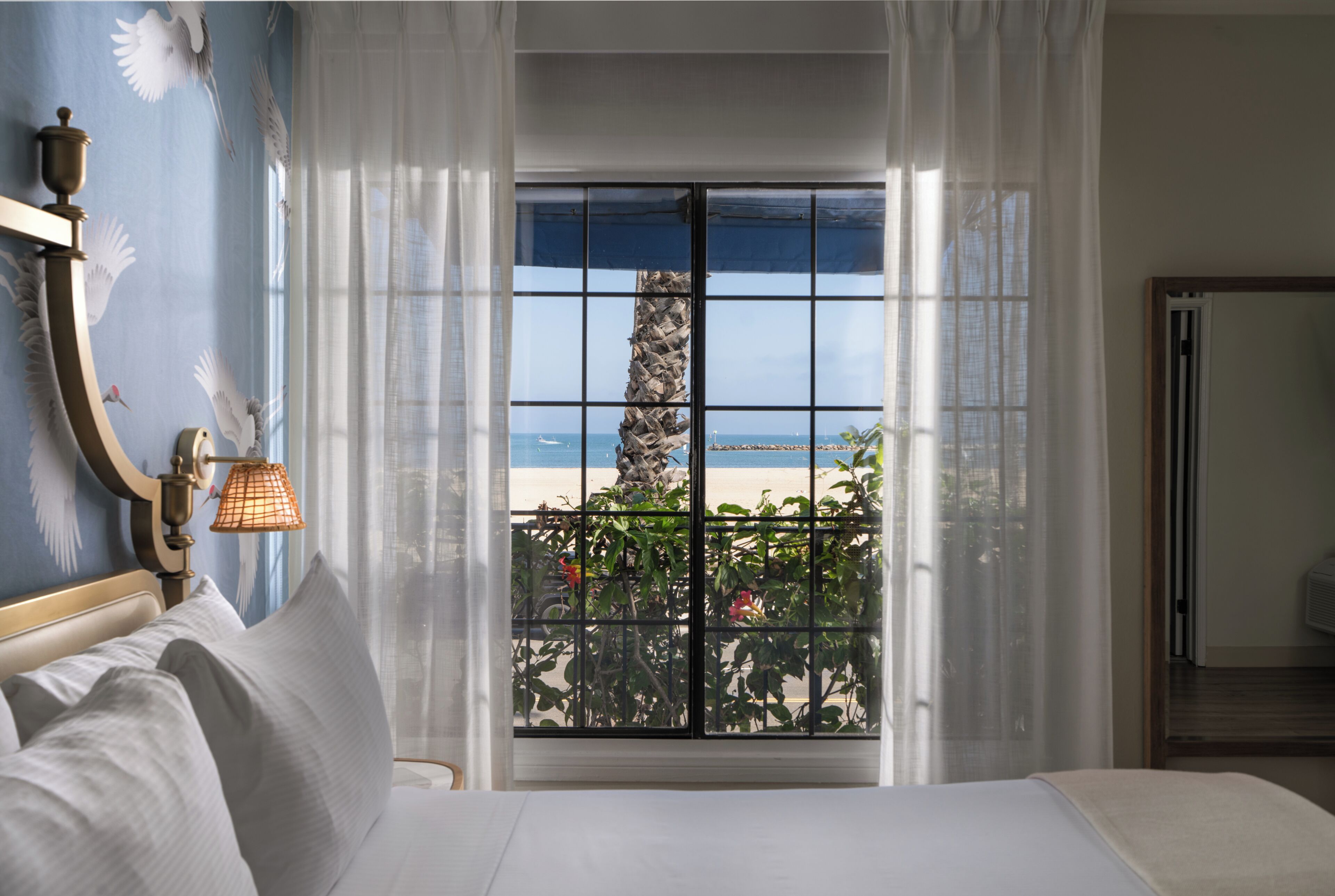 Una camera d'albergo con una grande finestra vista spiaggia, un letto e un comodino con una lampada.