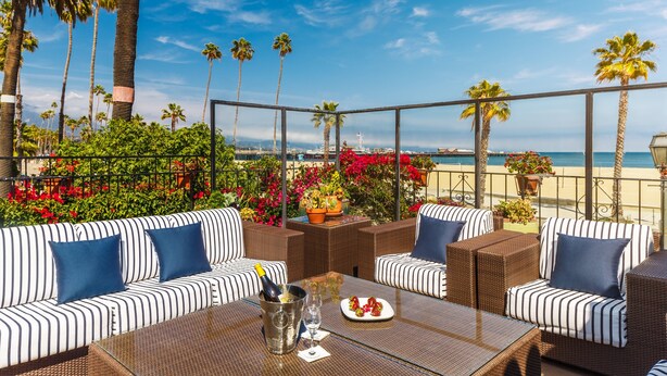 A patio with striped sofas and a table, overlooking a beach with palm trees and flowers.