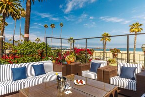 A patio with striped sofas and a table, overlooking a beach with palm trees and flowers.