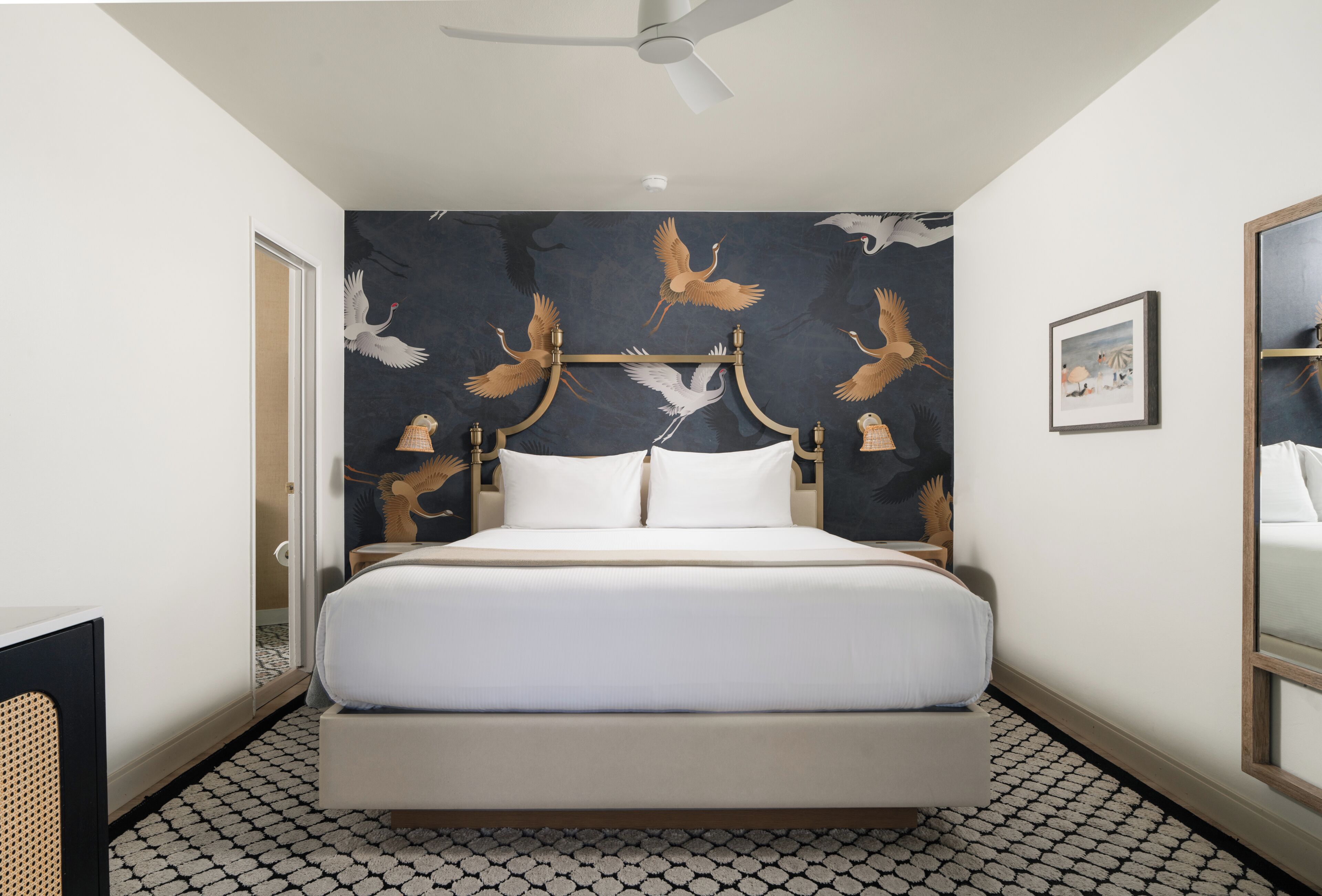 A hotel room with a bed, a ceiling fan, a wall with a bird mural, a framed picture, and a patterned floor.