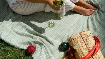 A person sitting on a checkered blanket outdoors, holding a glass of wine, with a wicker basket and a bottle nearby.