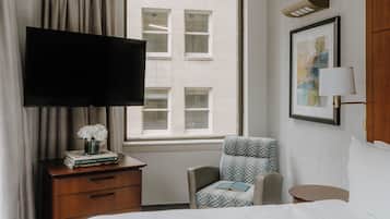 A hotel room with a flat-screen TV, a chair, a bedside table, a lamp, and a window with a view of a building.
