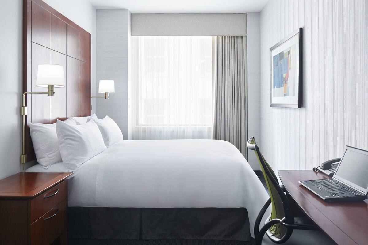 A hotel room with a bed, a desk with a laptop, a chair, a nightstand, and a window with curtains.