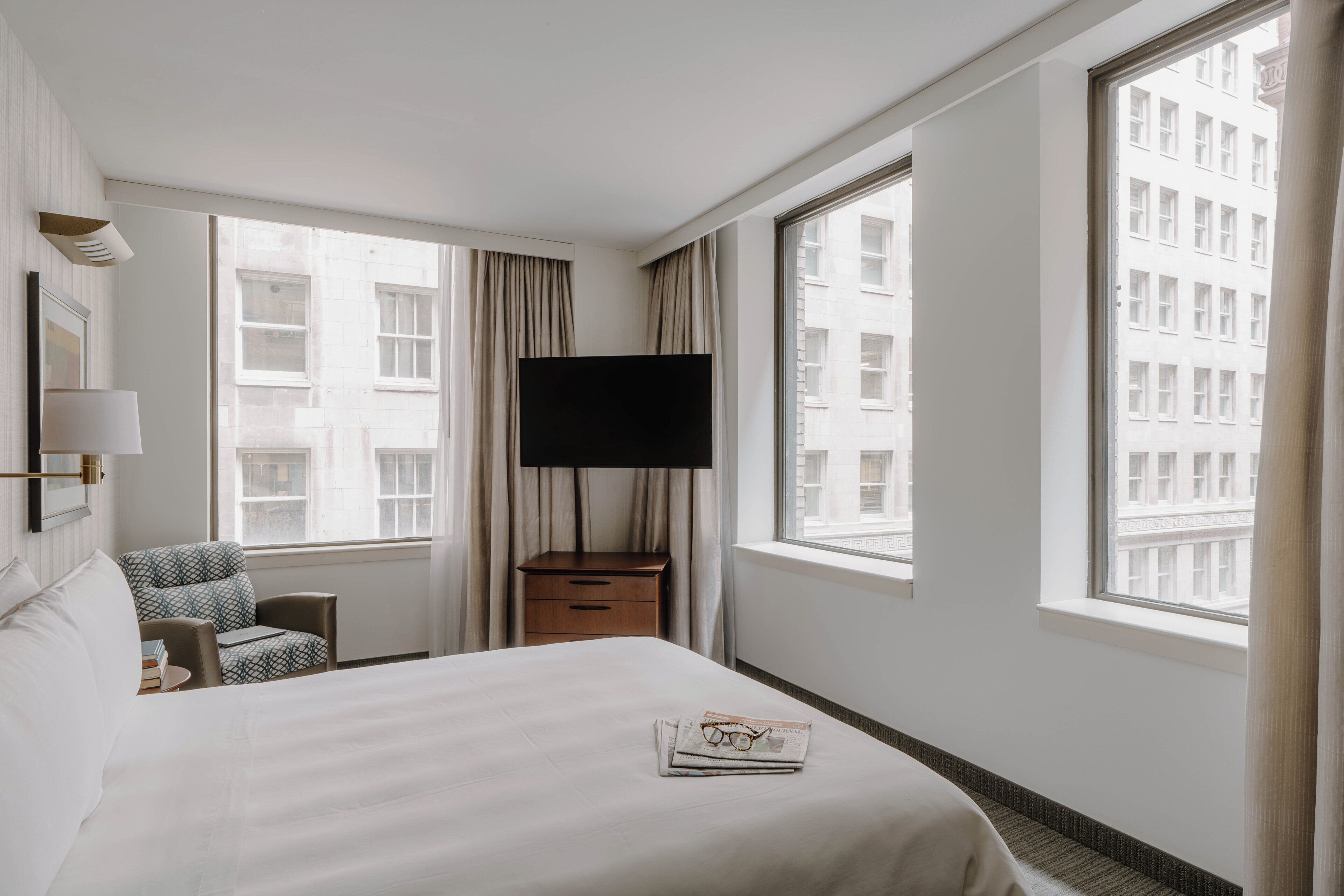 A hotel room with a bed, a TV, a chair, a nightstand, and large windows with a city view.