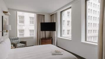 A hotel room with a bed, a TV, a chair, a nightstand, and large windows with a city view.