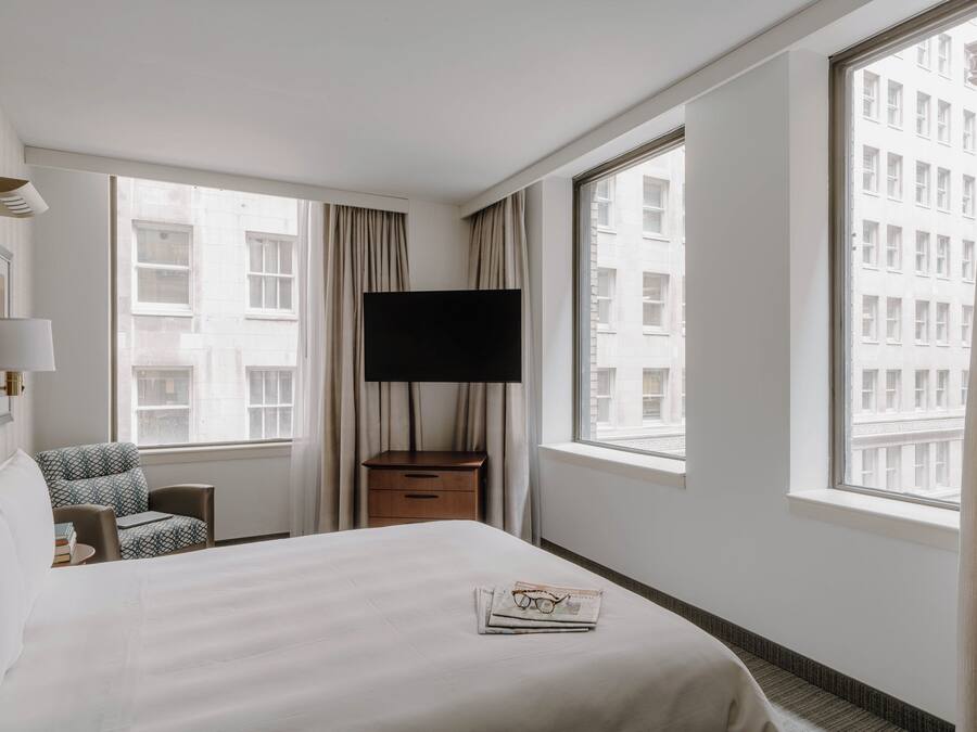 Habitación de hotel con cama, televisión, sillón, mesita de noche y ventanas amplias con vistas a la ciudad.