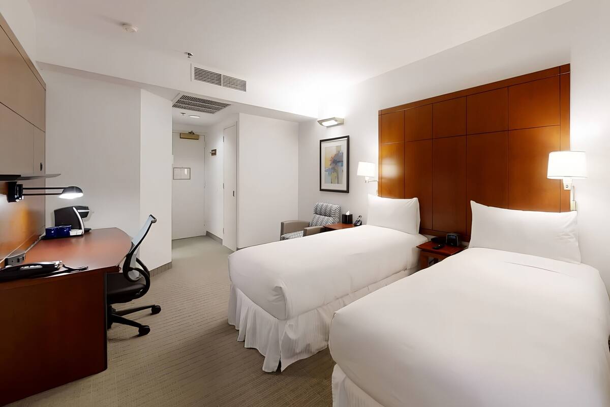 A hotel room with two beds, a desk with a lamp, a chair, a bedside table with a lamp, and a painting on the wall.