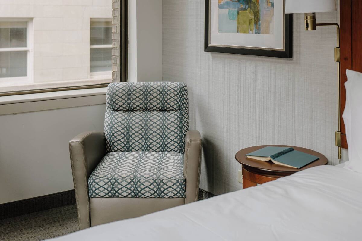A hotel room with a bed, a chair, a bedside table with a lamp, a framed abstract painting, and a window with a view of a building.