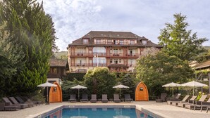 Un área de piscina con sillas de descanso y sombrillas, rodeada de árboles y un edificio grande con varios balcones.