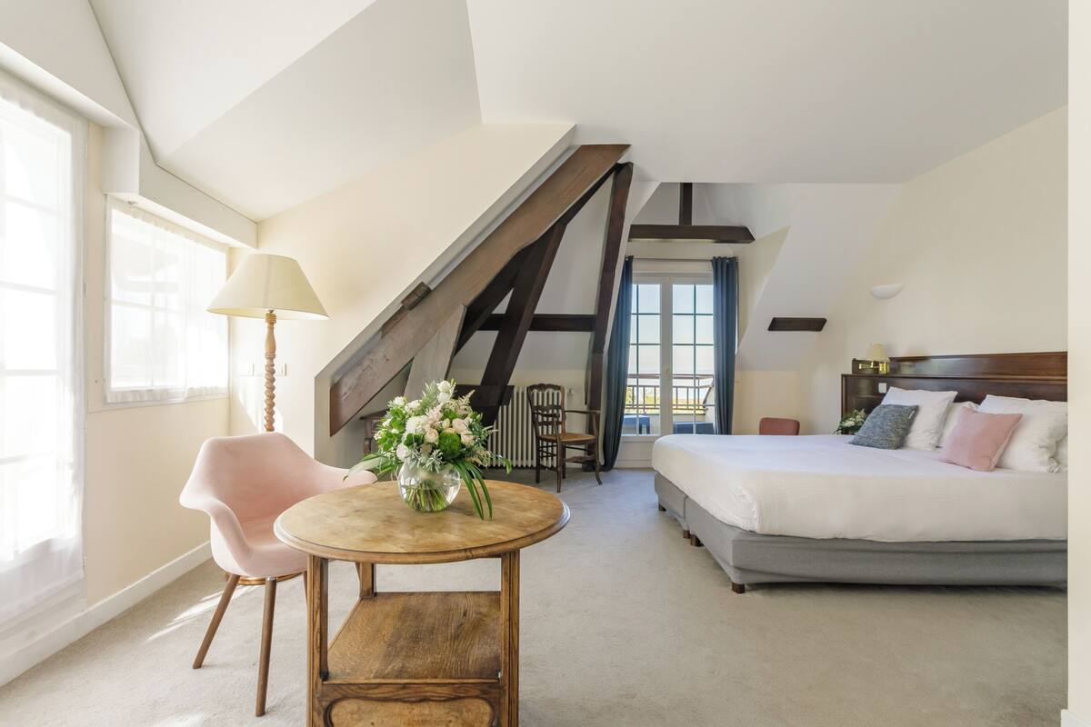 A bedroom with a bed, a round wooden table with a vase of flowers, a pink chair, and a staircase leading to an attic.