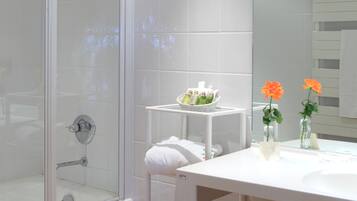Un baño moderno con cabina de ducha de vidrio, un lavamanos blanco con espejo y una mesita con artículos de aseo.