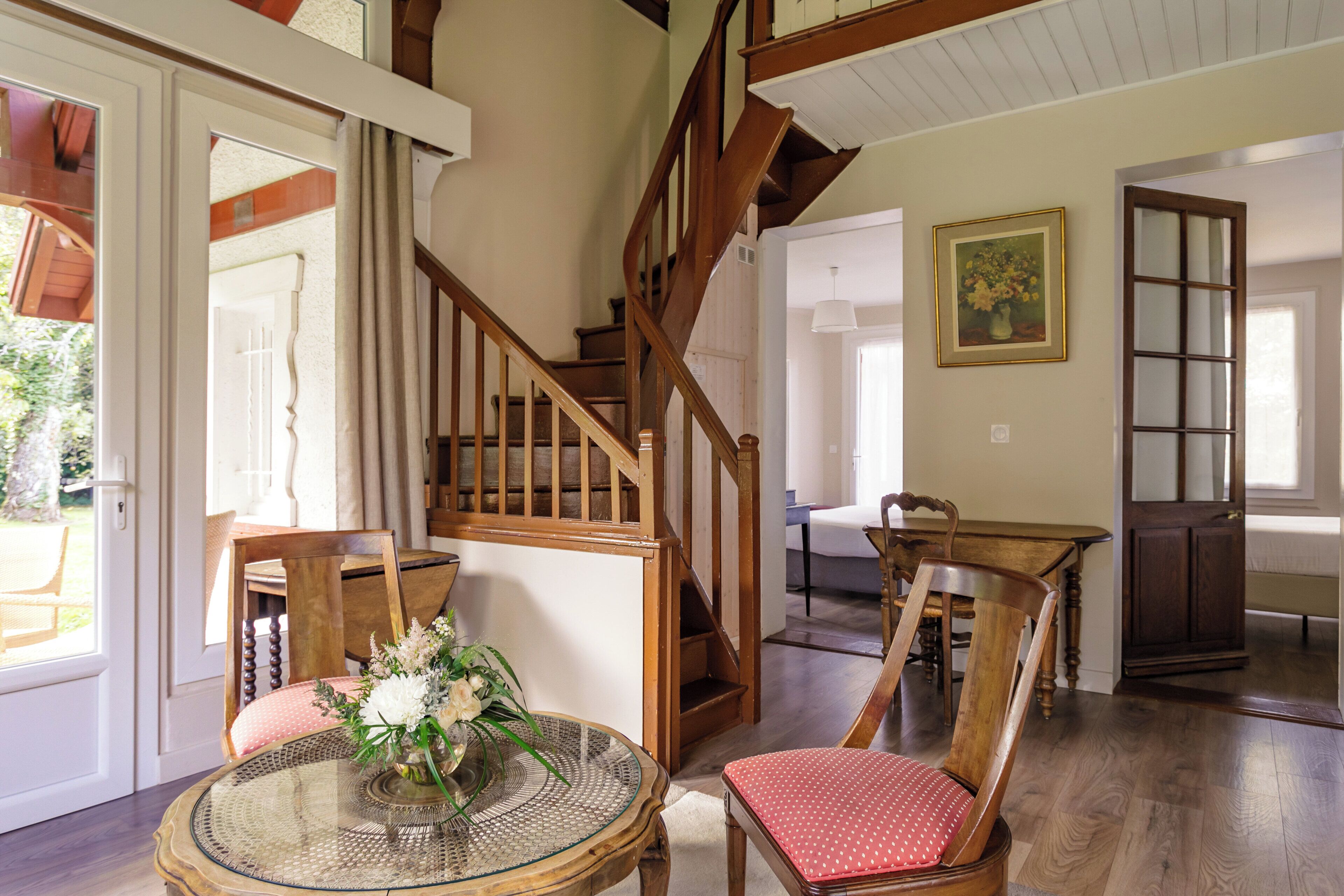A spacious room with a wooden staircase, a dining area with wooden chairs, and a round glass table with a vase of flowers.
