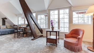 Une pièce de style « loft », avec un plafond haut, des poutres apparentes, une grande fenêtre, un canapé, un fauteuil, une table et une lampe.
