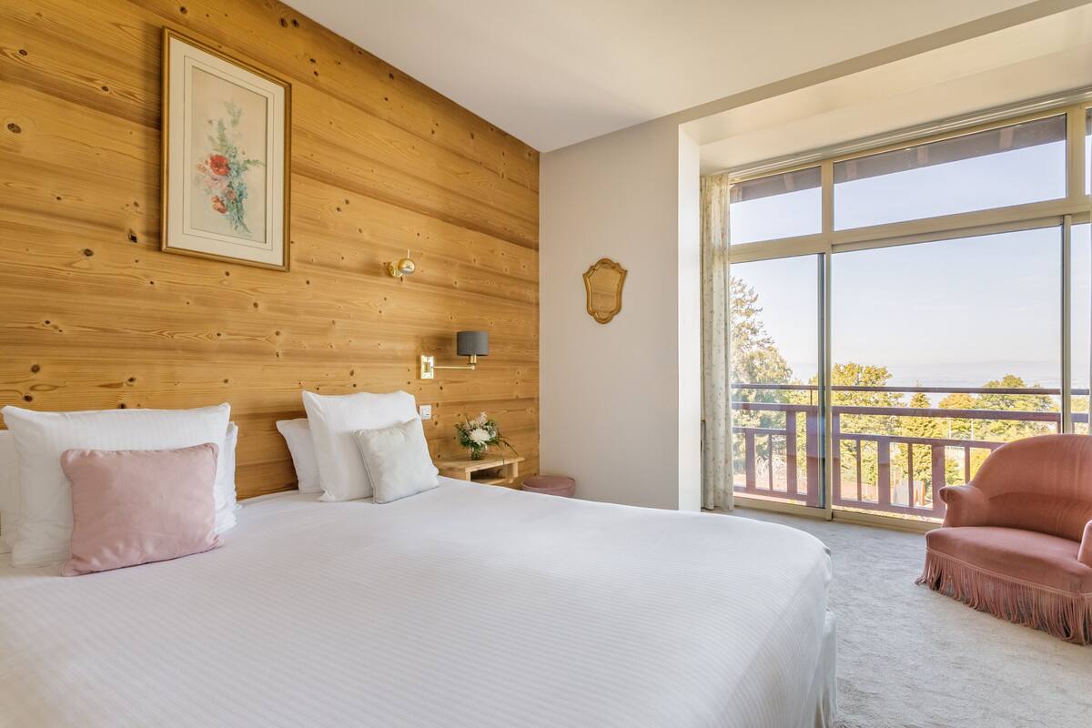 A bedroom with a large bed, a wooden headboard, a nightstand, a pink chair, and a balcony with a view.