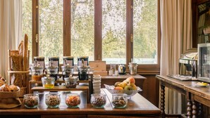 Un bufé de desayuno con diversos alimentos y condimentos, una máquina de café y vistas a árboles a través de amplias ventanas.