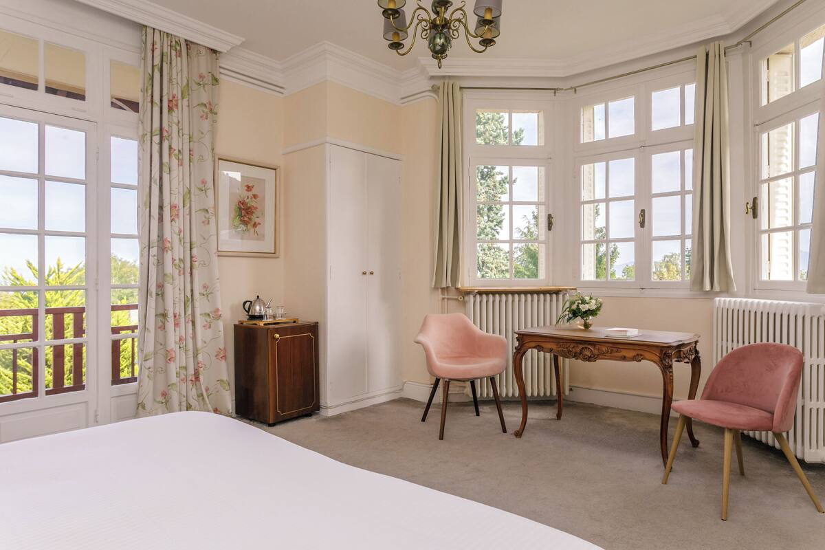 A room with a large bed, a desk with a chair, a radiator, and a window with curtains.