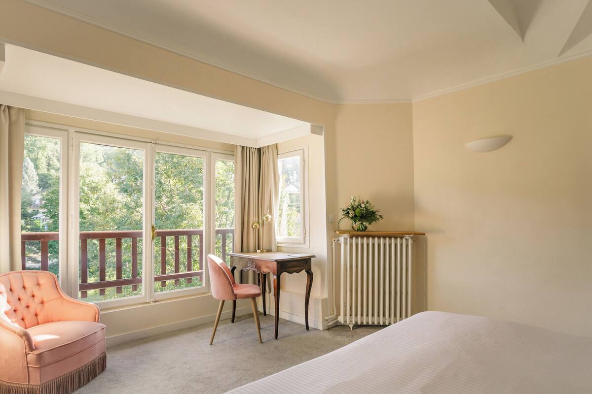 A bedroom with a bed, a desk, a chair, and large windows with a view of trees.