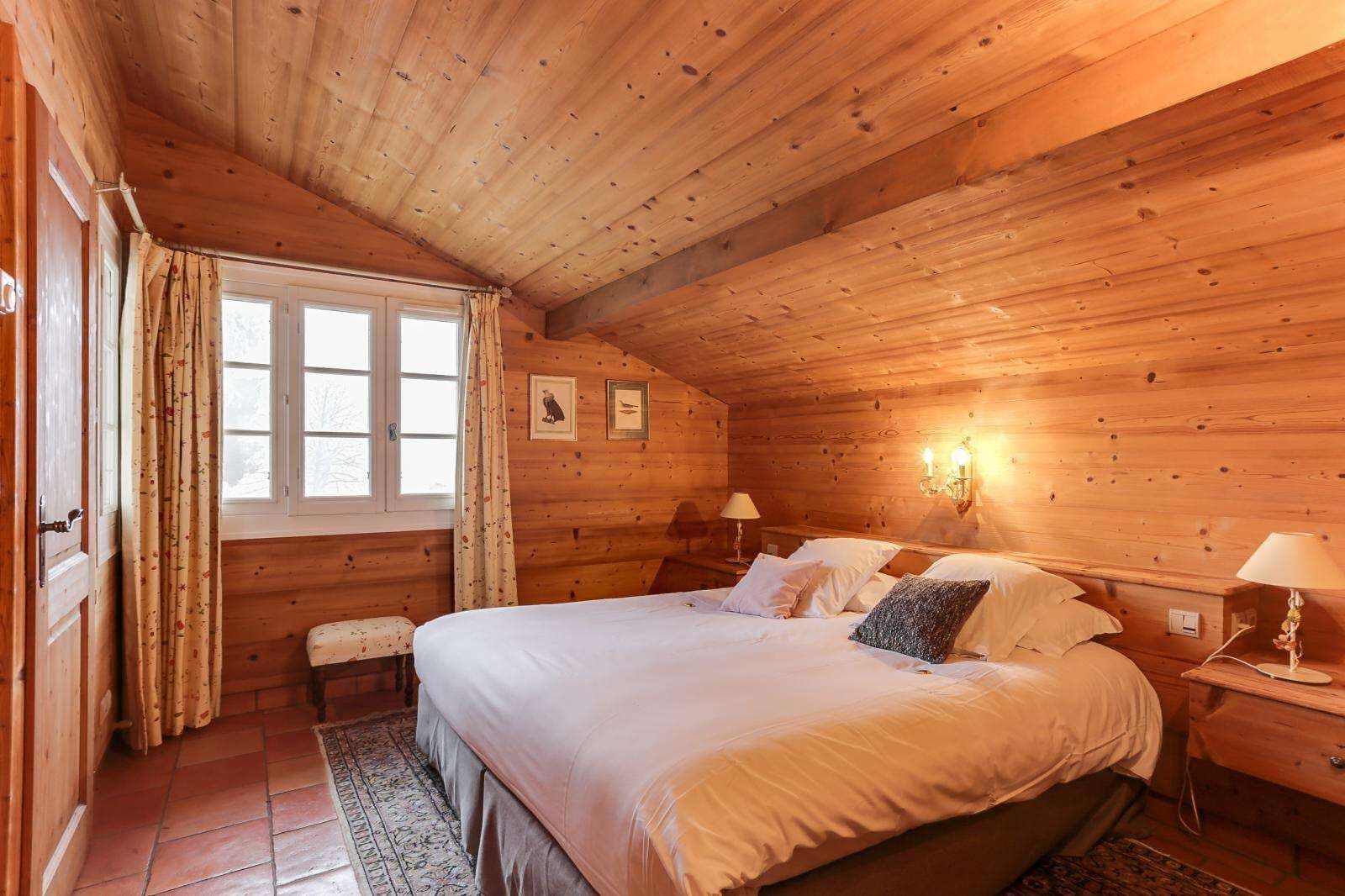 A wooden cabin bedroom with a large bed, two bedside tables, a bench, and a window with curtains.