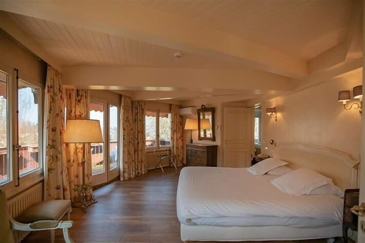 A bedroom with a large bed, a desk, and a chair. There are windows with curtains, a lamp, and wall sconces.