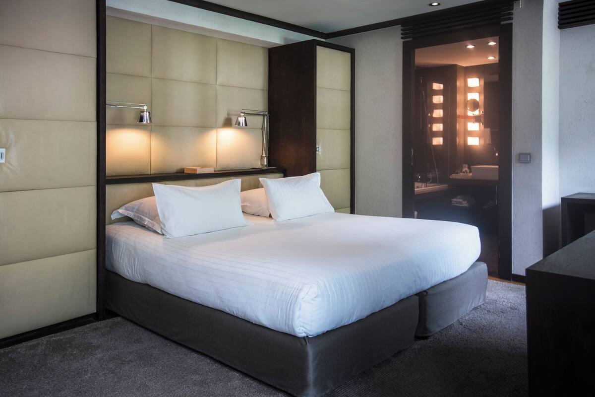 Ett modernt hotellrum med en stor säng, vadderat sänggavel, sänglampor och utsikt in i badrummet.