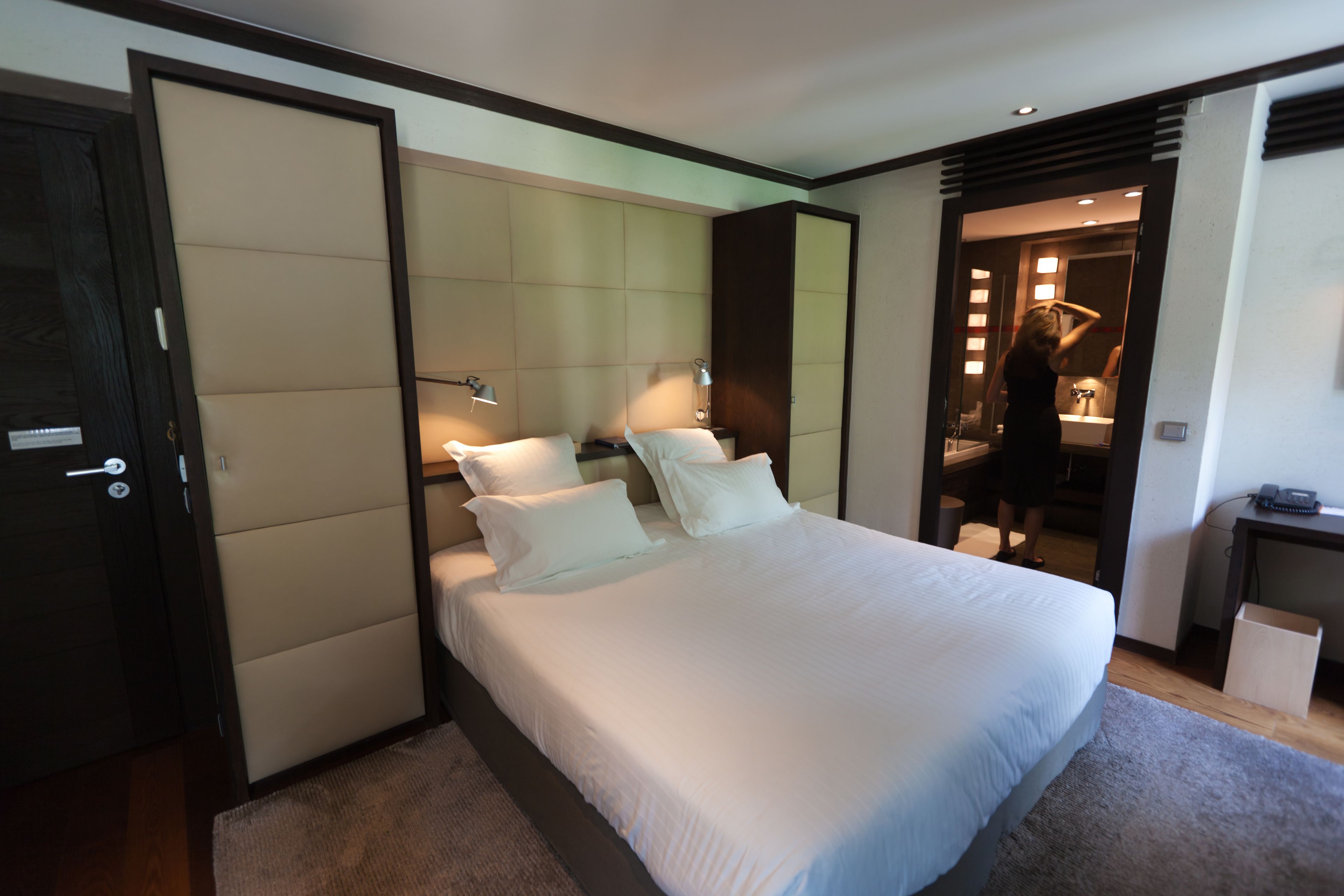 A modern hotel room with a large bed, a bedside table, a desk, and a bathroom visible through an open door.