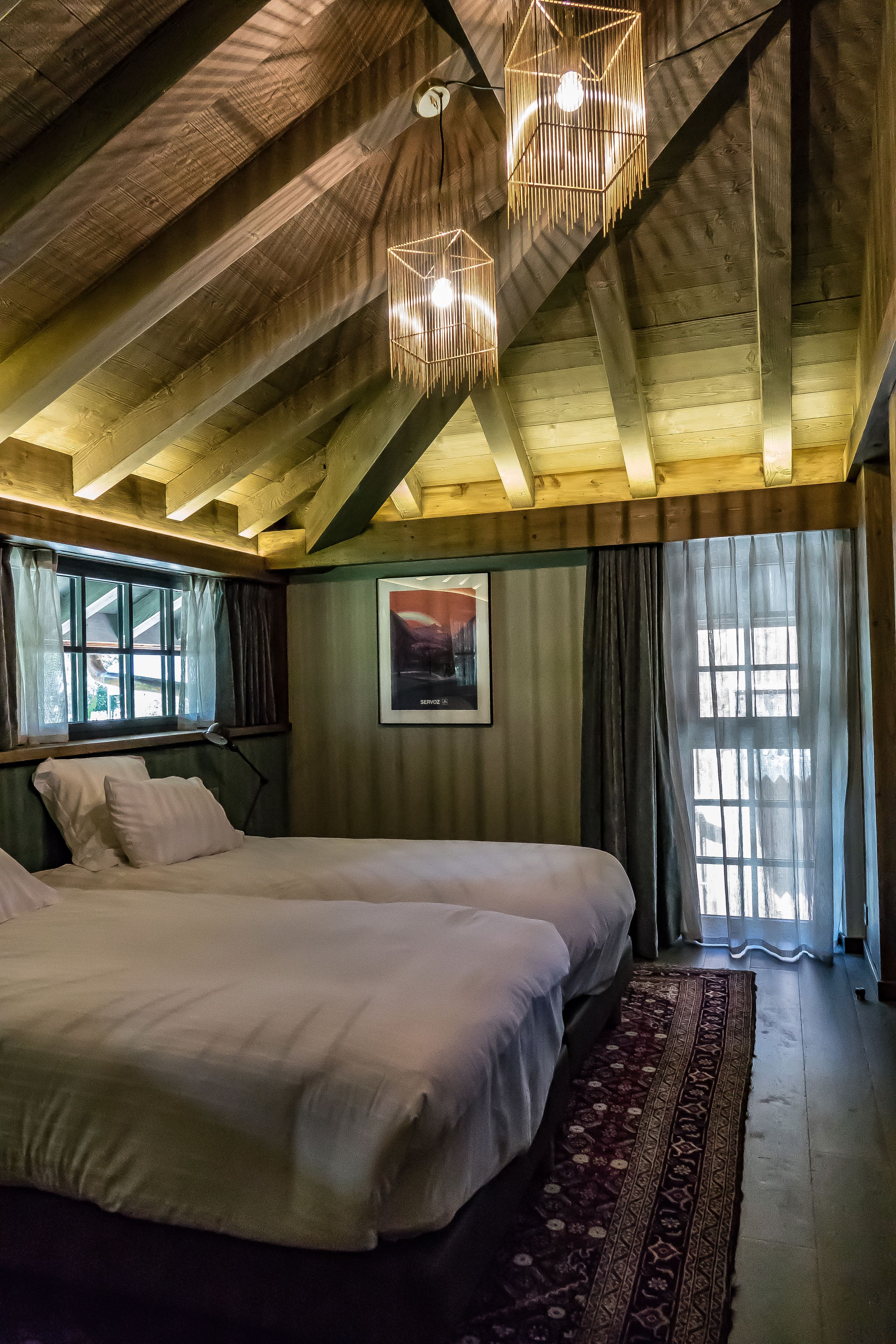 A bedroom with two beds, wooden beams on the ceiling, and a large window with curtains.