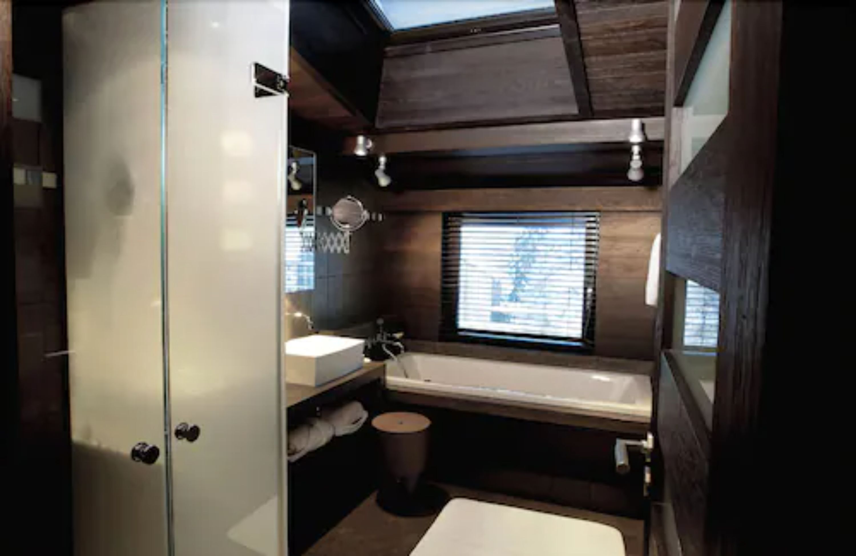 A compact bathroom with a glass shower enclosure, a bathtub, and a window with blinds.