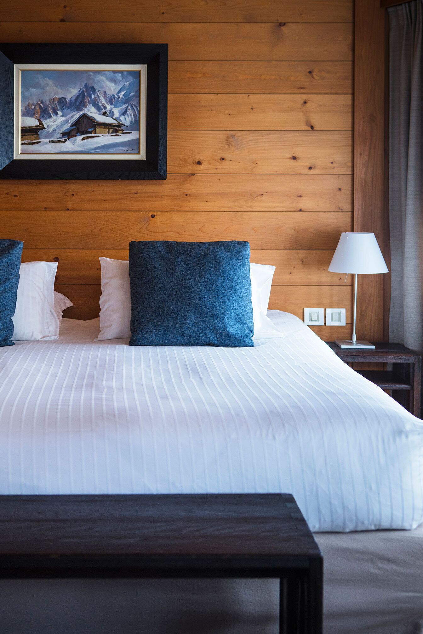 A hotel room with a large bed, wooden panel walls, a framed picture of a mountain, and a bedside lamp.