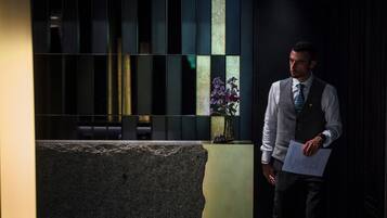 Un hombre con camisa y corbata de blanco, de pie junto a un muro de piedra con un jarrón de flores moradas.